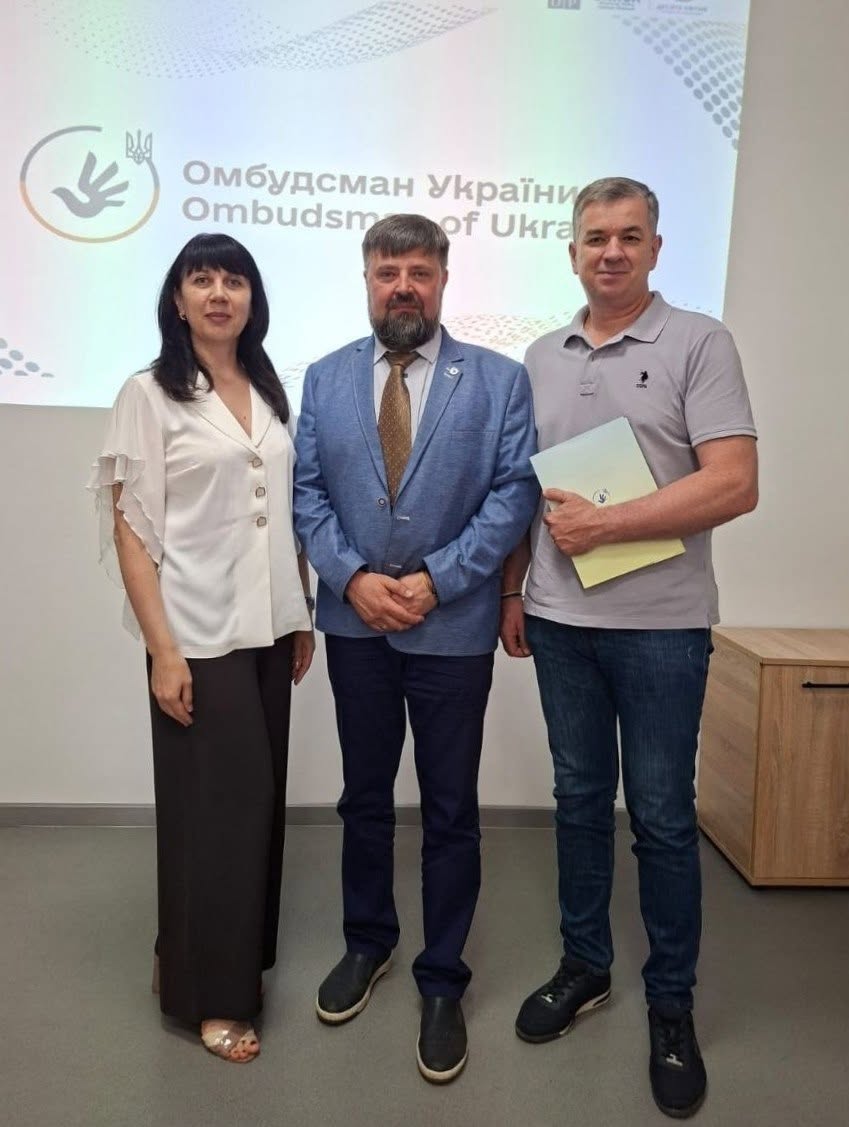 Три учасники зустрічі позують у повний зріст на тлі офіційного екрана.  Ліворуч стоїть жінка з темним волоссям і прямим чубчиком, одягнена у білу вільну блузку на ґудзиках та широкі чорні штани.  По центру стоїть бородатий чоловік у блакитному піджаку, білій сорочці, золотисто-коричневій краватці та темних штанях, який зчепив пальці рук перед собою.  Праворуч стоїть чоловік у сірій футболці-поло, синіх джинсах та чорних кросівках, який тримає в лівій руці закриту світлу папку з документами.  За їхніми спинами горить білий екран із великим написом: «Омбудсман України / Ombudsman of Ukra...». Праворуч на задньому плані помітно частину світлої дерев'яної офісної тумби.
