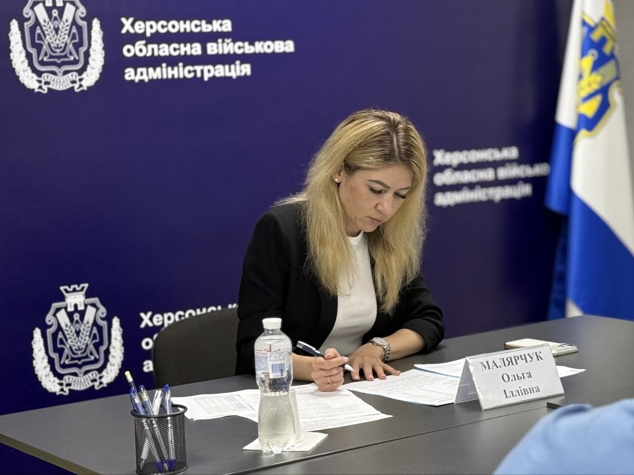 Заступниця начальника Херсонської обласної військової адміністрації Ольга Малярчук під час засідання. Жінка зі світлим волоссям одягнена у білу блузку та чорний піджак. Вона сидить за робочим столом, злегка нахиливши голову, і зосереджено підписує паперові документи кульковою ручкою. На її лівому зап'ясті — годинник на металевому ремінці.  Перед нею на столі встановлено табличку з написом «МАЛЯРЧУК Ольга Іллівна», стоїть прозора пластикова пляшка води «Моршинська» та підставка з ручками. Тло за її спиною — суцільний насичено-синій банер із білими офіційними гербами та повторюваним написом «Херсонська обласна військова адміністрація». Праворуч на задньому плані видно частину державного прапора України.