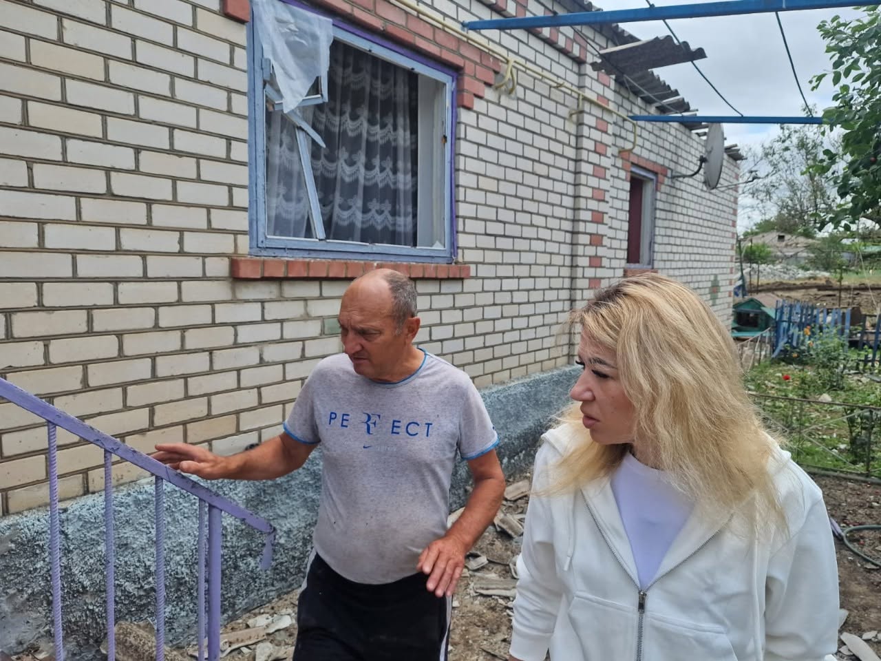 Вид на зруйновану вибухом житлову кімнату через дверний отвір. Повна шибок віконна рама вибита і лежить на ліжку, покритому пилом та дрібними осколками скла. На стінах кімнати наклеєні дитячі шпалери з малюнками тварин. Справа на тумбочці стоїть великий чорний телевізор, який дивом уцілів, а поруч із ним — коробки та речі. Через порожній проріз вікна видно стіну сусіднього будинку з білої цегли, на якій помітна глибока тріщина.