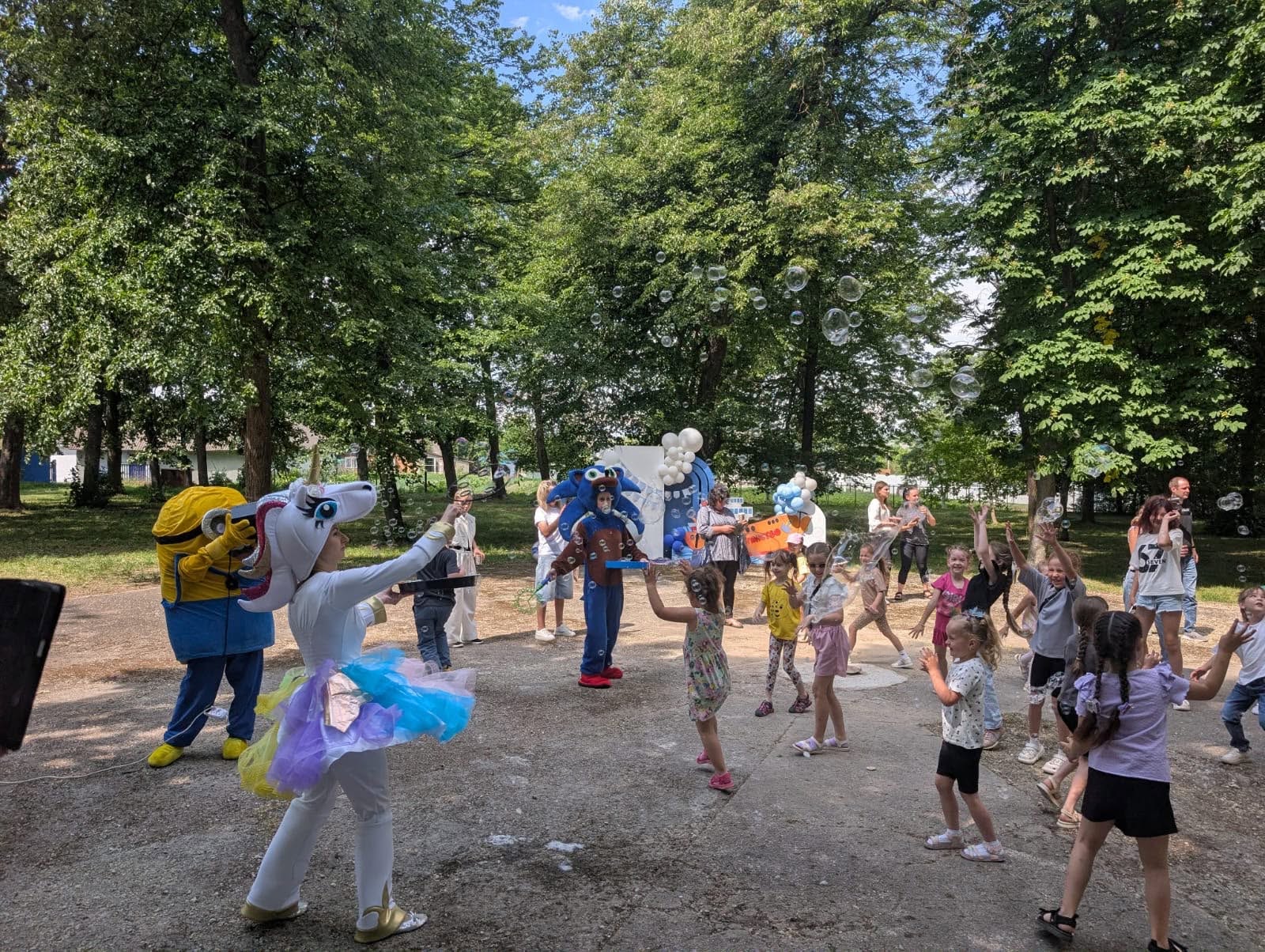 Розважальна програма для дітей у парку за участю аніматорів. На асфальтованому майданчику серед дерев бігають та стрибають близько п'ятнадцяти дітей різного віку. Перед ними виступають три аніматори в об'ємних ростових костюмах: ліворуч — жовтий Міньйон, по центру — синій персонаж (імовірно, Сонік), а на передньому плані ліворуч — аніматор у білому костюмі Єдинорога з пишною різнокольоровою фатиновою спідницею, який пускає мильні бульбашки. У повітрі навколо присутніх літає багато прозорих мильних бульбашок різного розміру. На задньому плані стоять дорослі та видно елементи святкового декору з повітряних кульок.
