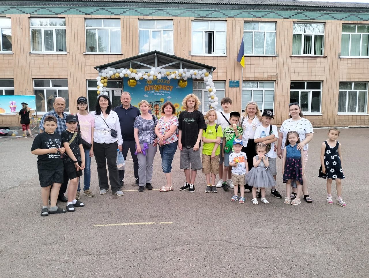 Групове фото учасників дитячого свята просто неба на подвір'ї навчального закладу. У два ряди позують близько двадцяти людей — діти різного віку, підлітки та дорослі. Усі одягнені в літній повсякденний одяг.  Позаду присутніх розташована двоповерхова цегляна будівля школи або дитячого садка з великими вікнами. Центральний вхід облаштований ґанком із навісом, який прикрашено великою аркою з білих та золотистих повітряних кульок. Під навісом встановлено яскравий блакитний святковий банер із малюнком соняшників та написом «2026 ЛІТОФЕСТ. Розпочнемо літо яскраво!». Праворуч від входу на стіні будівлі на флагштоці закріплено державний прапор України.  Учасники заходу посміхаються і дивляться в об'єктив камери; на передньому плані праворуч стоять найменші діти, серед яких хлопчик у блакитній кепці та дівчинка у пишній сірій сукні. На асфальтованому майданчику нанесено тонку жовту розмітку.