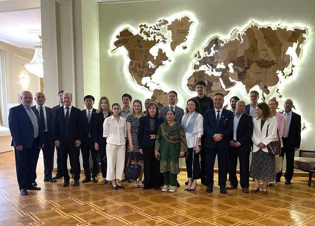 Спільне групове фото учасників міжнародної делегації та представників приймаючої сторони (близько двадцяти людей) у просторій залі. Чоловіки в офіційних ділових костюмах із краватками та жінки у сукнях, блузках і традиційному зеленому вбранні стоять в один ряд, посміхаючись у кадр. Вони позують на тлі великої світло-зеленої стіни, на якій встановлено масивний і яскраво підсвічений світлодіодами декоративний дерев'яний макет карти світу. Карта деталізована контурами материків і назвами країн англійською мовою (наприклад, Russia, China, Greenland). У залі покладено лакований дерев'яний паркет, ліворуч на другому плані видно кришталеву люстру.
