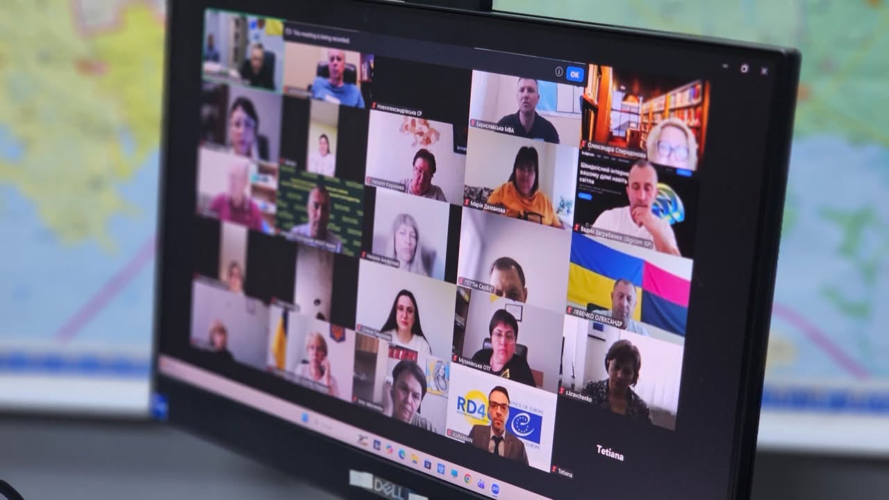 Екран чорного монітора бренду Dell під кутом, на якому відображається інтерфейс відеоконференції з великою кількістю вікон учасників онлайн-наради. Сітка трансляції містить понад двадцять інтерактивних віконець посадовців та представників громад.  Серед підписаних вікон видно: «Бериславська МВА», «Марія Денисова», «Лещенко Олександр» (на тлі синьо-жовтого та червоно-чорного прапорів), «Музиківська ОТГ», «Новоолександрівська СР». У нижній частині екрана, у вікні з емблемою Ради Європи та написом «RD4», транслюється чоловік в окулярах та костюмі. На розмитому задньому плані ліворуч помітно частину настінної географічної карти.