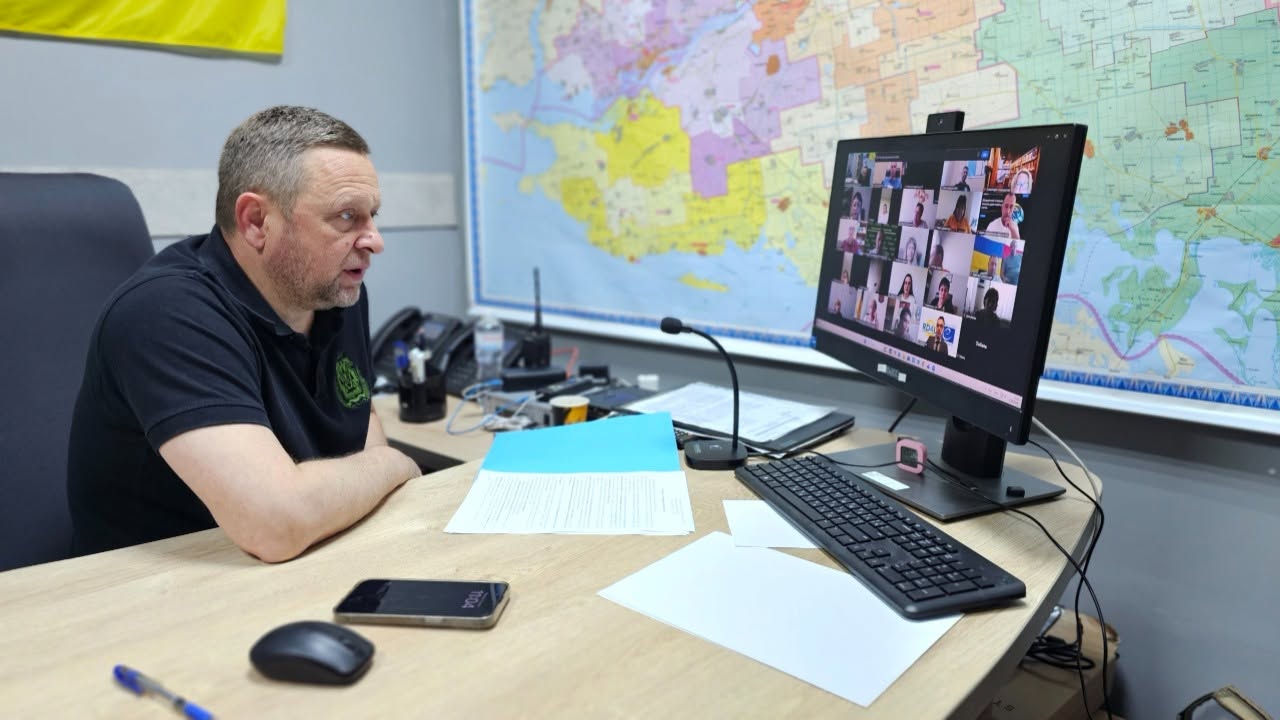 Перший заступник начальника Херсонської обласної військової адміністрації Дмитро Бутрій бере участь у робочій онлайн-нараді, сидячи за своїм робочим столом у кабінеті. Чоловік зображений у профіль ліворуч, він одягнений у чорну футболку-поло з зеленим логотипом на грудях та дивиться на екран комп'ютера, ведучи діалог.  На світлому дерев'яному столі перед ним розкладено папери, лежить блакитна папка з документами, мобільний телефон, комп'ютерна миша, чорна клавіатура та встановлено настільний мікрофон на гнучкій ніжці. На моніторі відображається сітка відеоконференції з учасниками. Увесь задній план за монітором займає велика настінна кольорова карта Херсонської області зі схематичним поділом на громади. У лівому верхньому кутку видно частину державного прапора України.