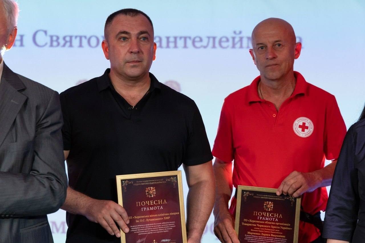 Великий план двох чоловіків-лауреатів на сцені, які тримають у руках свої відзнаки.  Ліворуч чоловік у чорній футболці-поло тримає в руках «ПОЧЕСНУ ГРАМОТУ», видану від імені Поважної ради Відзнаки за професіоналізм та милосердя «Орден Святого Пантелеймона» для КНП «Херсонська міська клінічна лікарня ім. О.С. Лучанського» ХМР.  Праворуч від нього стоїть лисий чоловік у червоній футболці-поло з білим круглим логотипом Товариства Червоного Хреста України на грудях. Він тримає аналогічну грамоту, виписану на ім'я Марченко Лариси Вацлавівни від ГО «Херсонська обласна організація Товариства Червоного Хреста України».  На задньому плані видно частину білого екрана з розмитим текстом.