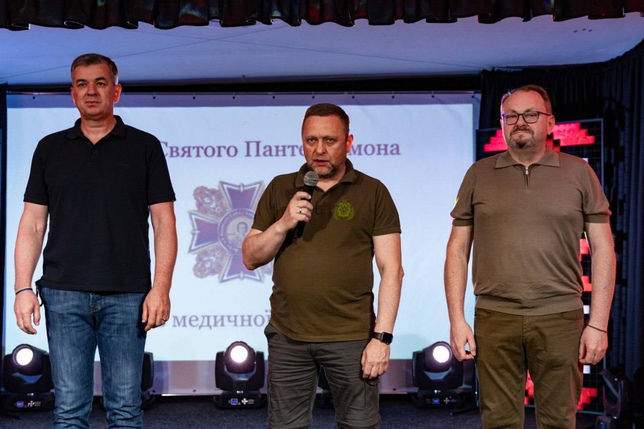 Три чоловіки стоять у ряд на сцені під час виступів.  Ліворуч стоїть чоловік у чорній футболці та синіх джинсах.  По центру з мікрофоном у руках виступає заступник начальника Херсонської ОВА Дмитро Бутрій.  Праворуч від нього стоїть чоловік в окулярах та оливковій футболці-поло з блискавкою на комірі.  На великому білому екрані за їхніми спинами відображається презентаційний слайд із зображенням великого фіолетового хреста — емблеми Ордена Святого Пантелеймона — та текстовим підписом «...святого Панте...мона ...медичної». Нижню частину сцени підсвічують два увімкнені сценічні прожектори.