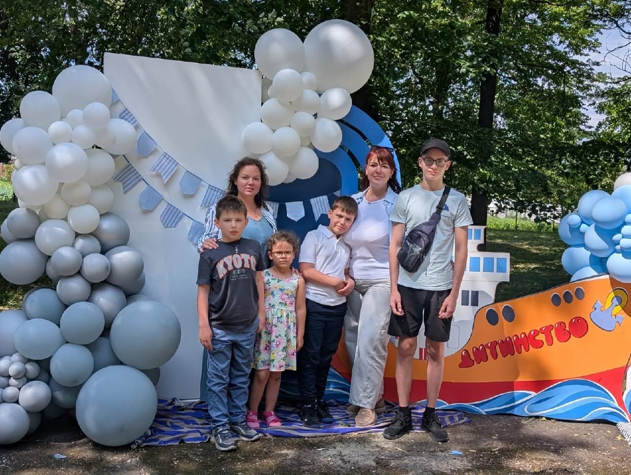 Групове фото сімох учасників свята — трьох жінок, трьох хлопчиків та однієї дівчинки — біля тематичної фотозони в парку. Вони стоять на синьо-білому покривалі, постеленому на землі. Фотозона оформлена у морському стилі: білий вертикальний щит прикрашений гірляндами з блакитних прапорців та великими арками з білих, сірих і блакитних повітряних кульок. Праворуч розташований яскравий помаранчевий декоративний макет корабля з хвилями біля основи та червоним написом «Дитинство». Усі присутні одягнені в літній повсякденний одяг і дивляться в кадр. Позаду них височіють зелені дерева парку.