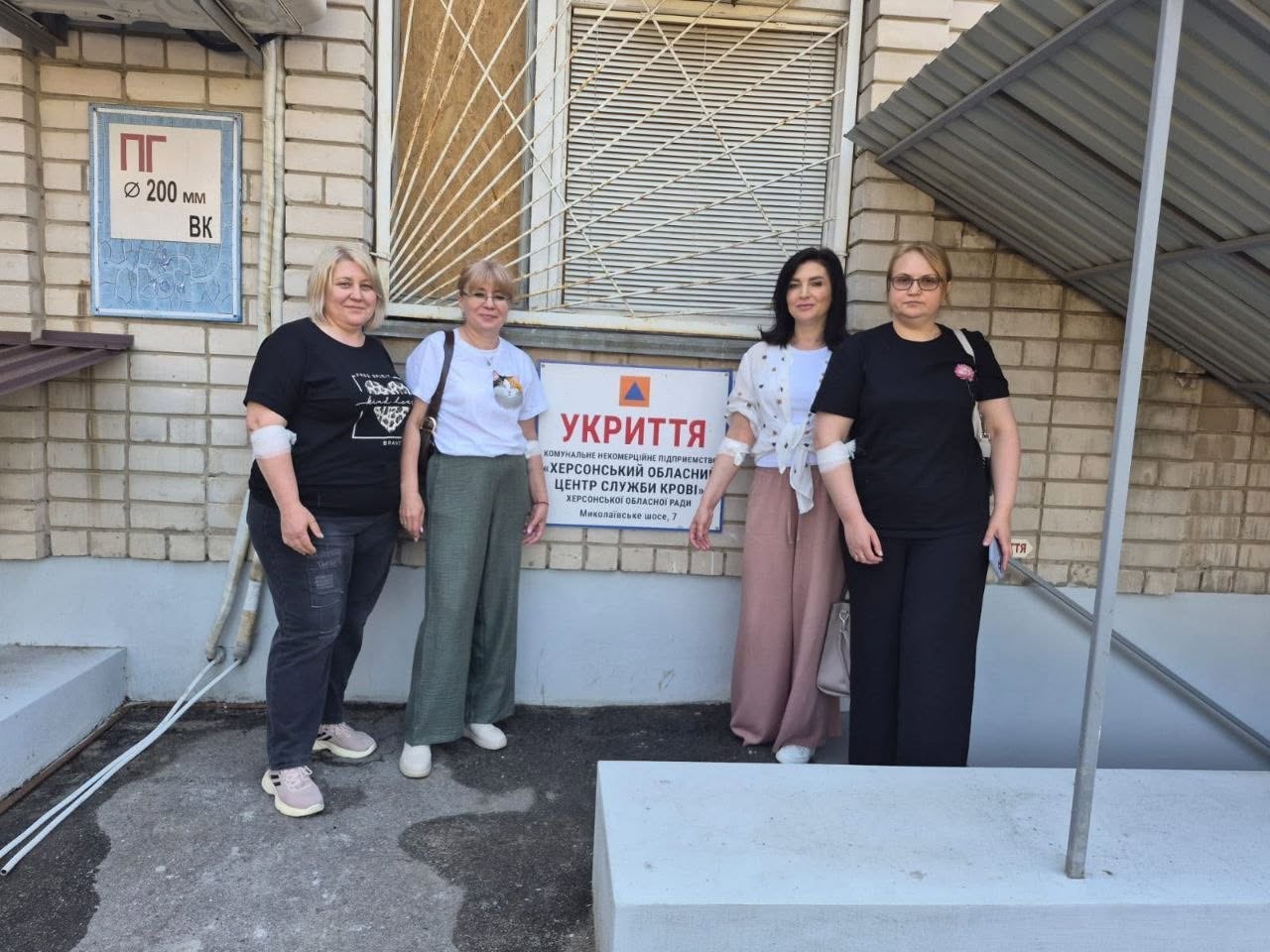 Чотири жінки-донорки позують для спільного фото на тлі цегляної стіни будівлі Херсонського обласного центру служби крові. Вони стоять двома парами по обидва боки від білої інформаційної таблички з написом «УКРИТТЯ. КНП «Херсонський обласний центр служби крові» ХО State Council, Миколаївське шосе, 7». У трьох жінок на ліктях накладено еластичні білі медичні пов'язки після процедури здачі крові. Дві відвідувачки одягнені в чорні футболки, одна — у білу з котиком, ще одна — у білу з візерунком та рожеві штані. На задньому плані видно заґратоване вікно та частину металевого навісу.
