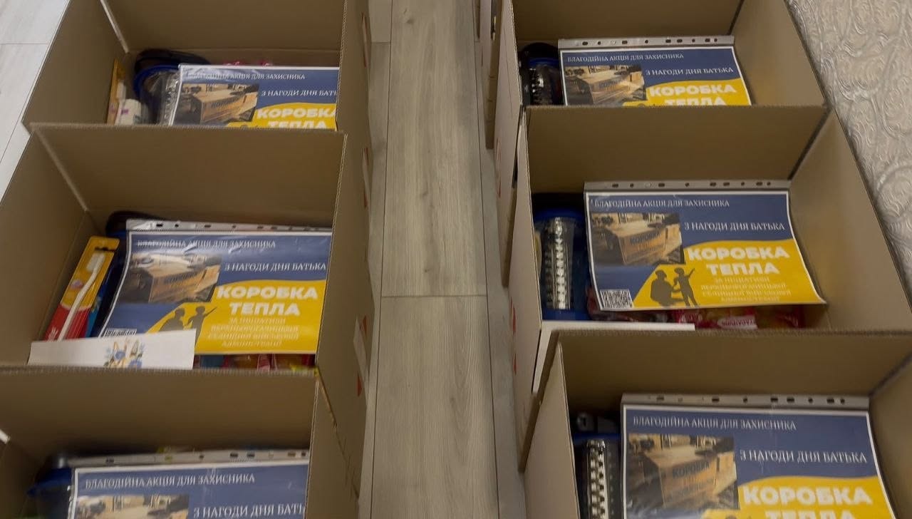 Велика кількість закритих однакових картонних коробок з гуманітарною допомогою, які акуратно складені у чотири ряди на підлозі біля світлої стіни. На кожну коробку наклеєно велику фірмову етикетку з малюнком червоної вантажівки, з кузова якої вилітає безліч червоних сердечок.  На лівій частині етикетки зображено сільський пейзаж із річкою та написом «ВЕРХНІЙ РОГАЧИК». Також на коробках присутній невеликий напис «25 років. Доставка любові» та надруковано квадратний QR-код для зчитування інформації.