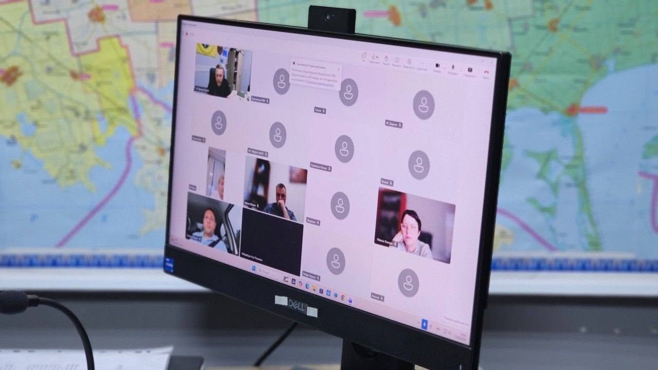 Екран чорного комп'ютерного монітора бренду Dell, знятий під кутом, на якому відображається інтерфейс відеоконференції. На екрані видно сітку вікон учасників онлайн-наради: кілька віконець транслюють відео (зокрема видно чоловіка в автомобілі та жінку у світлому одязі), тоді як більшість інших відображаються у вигляді сірих круглих іконок профілів без увімкнених камер. По центру екрана спливло невелике системне вікно сповіщення. На розмитому задньому плані за монітором розташована велика настінна географічна карта з різнокольоровими зонами та береговою лінією.