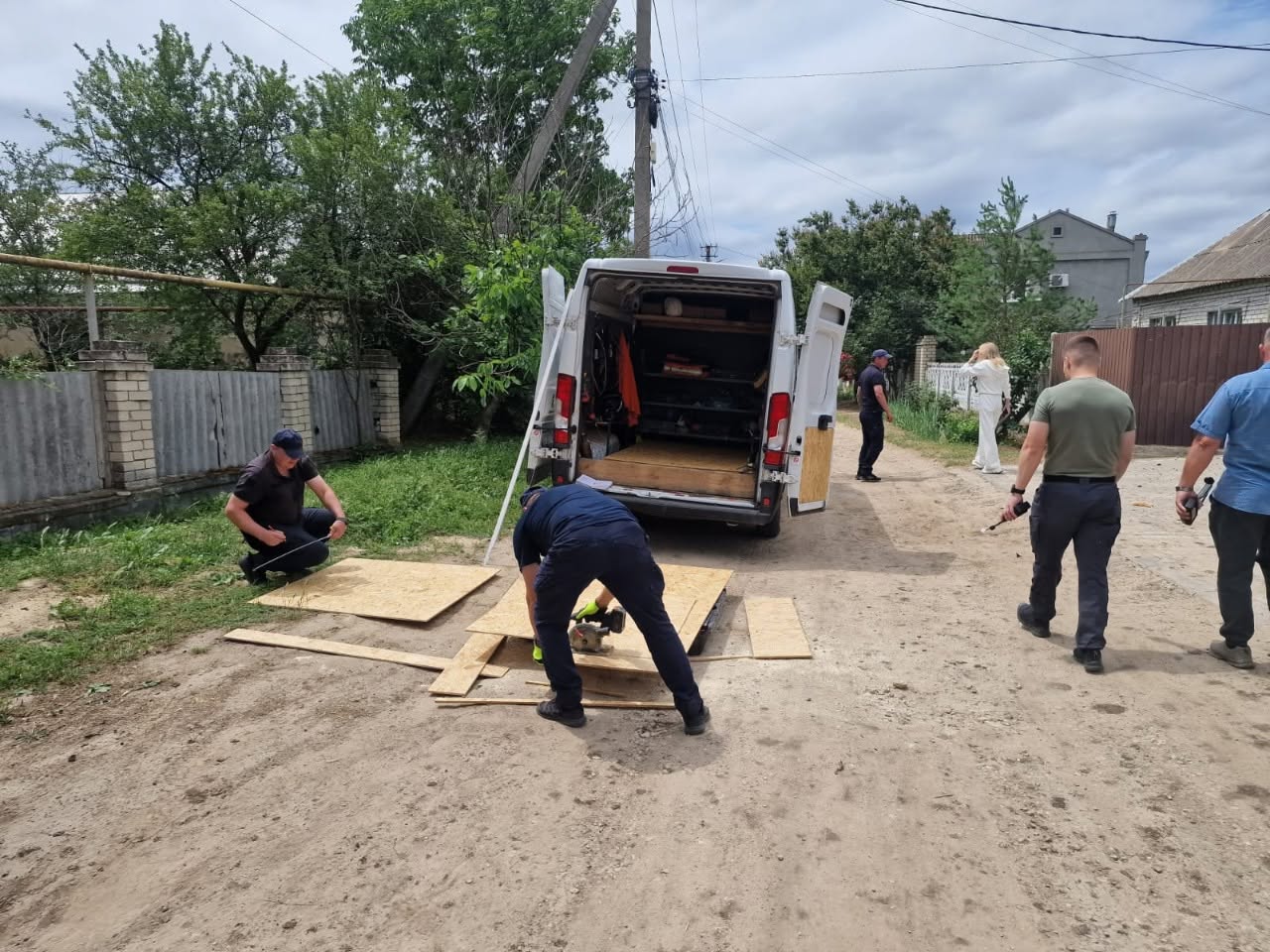 Ольга Малярчук у білому костюмі спілкується біля фігурного бетонного паркану з місцевою мешканкою, яка постраждала від обстрілу. Літня жінка у зеленій футболці та хустці на голові сперлася на паркан, тримаючи в руці робочі рукавиці, і з важким емоційним виразом обличчя розповідає про пережите. Заступниця начальника ОВА уважно вислуховує жінку. На задньому плані видно синє металеве подвірне приналежність, складений ракушняк та силует іншої людини в камуфляжній футболці.