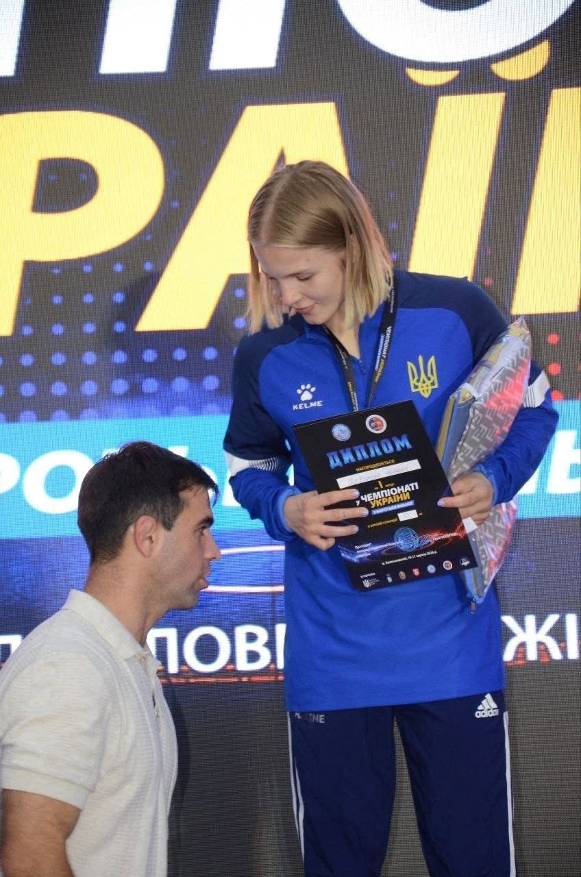 Великий план моменту нагородження. На п'єдесталі стоїть світловолоса дівчина-спортсменка в офіційному синьому спортивному костюмі збірної України, на грудях якого розміщено герб (тризуб) та логотип бренду Kelme. Вона схилила голову, розглядаючи чорний диплом у своїх руках із написом «ДИПЛОМ за 1 місце у ЧЕМПІОНАТІ УКРАЇНИ з боротьби вільної». Під пахвою лівої руки вона тримає загорнутий у плівку подарунок чи кубок. Зліва знизу у профіль зафіксовано чоловіка у світлій футболці-поло, який дивиться на спортсменку. Тлом слугує великий світлодіодний екран, де видно фрагменти написів «...РАЇН...», «...пові...» та «...ЖІ...».