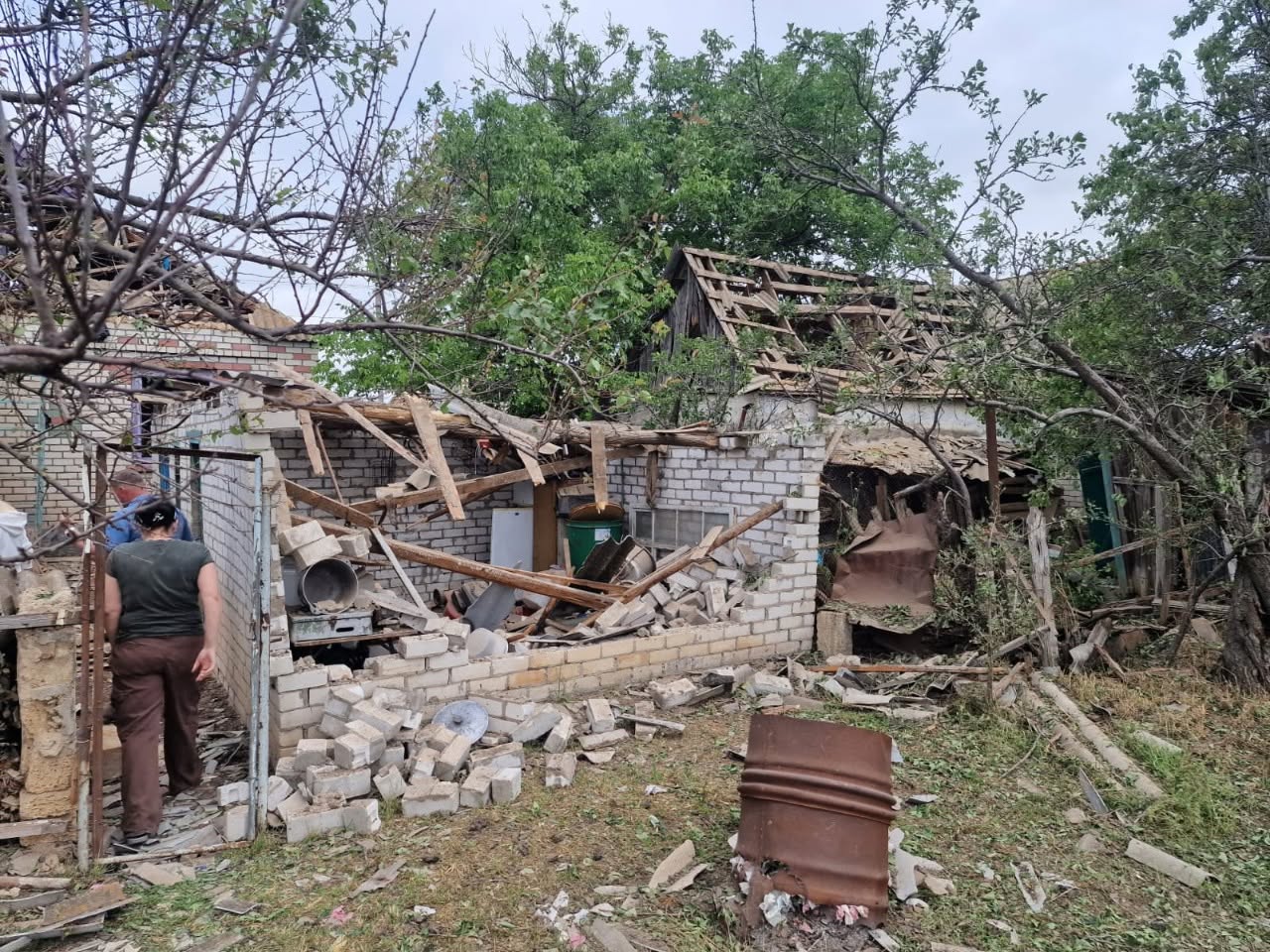 Чоловік у світло-сірій футболці та темних джинсах стоїть у центрі величезної та глибокої вирви від вибуху ворожого снаряда, демонструючи її масштаб. Вирва утворилася посеред городу чи поля, навколо видно переорану вибуховою хвилею землю та брили ґрунту. На задньому плані кадру, на межі ділянки, стоїть стара вантажівка з фургоном, видно пошкоджені одноповерхові житлові будинки та зелені дерева. Погода хмарна.