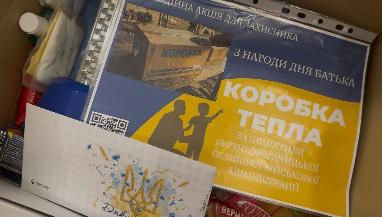 Вид згори на вміст відкритої картонної коробки з гуманітарною допомогою. Всередині пакунка видно продукти, гігієнічні засоби та святкову листівку від Укрпошти із зображенням золотавих колосків пшениці, волошок та тризуба.  Поверх речей лежить яскрава інформаційна листівка у пластиковому файлі. На ній великими білими літерами на жовто-синьому тлі написано: «КОРОБКА ТЕПЛА ЗА ІНІЦІАТИВИ ВЕРХНЬОРОГАЧИЦЬКОЇ СЕЛИЩНОЇ ВІЙСЬКОВОЇ АДМІНІСТРАЦІЇ». У верхній частині листівки міститься напис: «БЛАГОДІЙНА АКЦІЯ ДЛЯ ЗАХИСНИКА З НАГОДИ ДНЯ БАТЬКА», а в центрі зображено силует чоловіка та дитини, яка вказує рукою вперед.