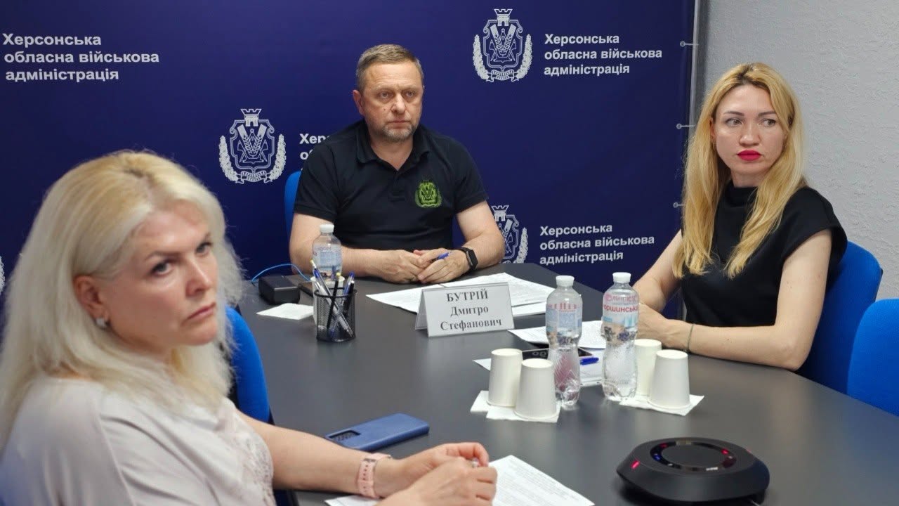 Перший заступник начальника Херсонської ОВА Дмитро Бутрій та представниці профільних підрозділів беруть участь у засіданні за робочим столом сірого кольору.  Дмитро Бутрій сидить по центру в анфас, він одягнений у чорну футболку-поло з невеликим зеленим гербом на грудях. Перед ним встановлено іменну табличку «БУТРІЙ Дмитро Стефанович», лежать робочі документи та стоїть підставка з ручками.  Ліворуч на передньому плані у профіль зафіксована жінка зі світлим волоссям у світлій блузці.  Праворуч за столом сидітиме інша учасниця наради — блондинка у чорній блузці з яскравою помадою.  На столі перед присутніми розставлені пластикові пляшки з водою, паперові стаканчики, лежить мобільний телефон та розміщено круглий чорний пристрій гучного зв'язку (спікерфон). Тло за спинами посадовців оформлене суцільним темно-синім пресволлом із повторюваним білим гербом області та офіційним написом «Херсонська обласна військова адміністрація». Права частина кадру межує із сірою стіною кабінету.