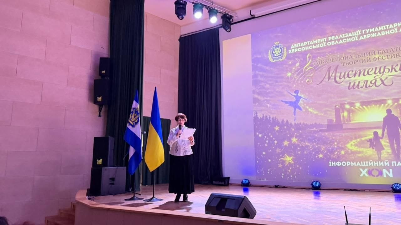 Ведуча заходу виступає на сцені на тлі великого мультимедійного екрана. Жінка в окулярах, білій вишитій блузці та довгій чорній спідниці стоїть по центру сцени, тримаючи в руці мікрофон та папери з текстом промови.  Ліворуч від неї на підставках встановлено два державні прапори України: один повністю синьо-жовтий, інший — синій із золотистим гербом (тризубом). На великому екрані ззаду горить фіолетово-рожева заставка фестивалю із зображенням силуету балерини, що танцює, нотного стану та офіційними написами: «ДЕПАРТАМЕНТ РЕАЛІЗАЦІЇ ГУМАНІТАРНОЇ ПОЛІТИКИ ХЕРСОНСЬКОЇ ОБЛАСНОЇ ДЕРЖАВНОЇ АДМІНІСТРАЦІЇ» та «МІЖРЕГІОНАЛЬНИЙ БАГАТОЖАНРОВИЙ ТВОРЧИЙ ФЕСТИВАЛЬ «Мистецький шлях»». Сцена обладнана професійними підвісними та підлоговими прожекторами.