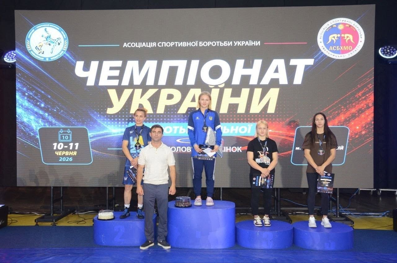 Загальний план церемонії нагородження переможців на чемпіонаті України з вільної боротьби серед жінок. На синьому трирівневому круглом п'єдесталі позують чотири призерки та тренер: На першій сходинці (по центру) стоїть чемпіонка в синій спортивній кофті збірної України, тримаючи в руках нагороду. На другій сходинці (ліворуч) стоїть дівчина у синій футболці з медаллю на шиї. Попереду неї на підлозі стоїть чоловік у білій футболці-поло та сірих штанях. На третій сходинці (праворуч) стоїть спортсменка в чорній футболці з медаллю. Поруч із нею (крайня праворуч, поза п'єдесталом) стоїть ще одна призерка у коричневій футболці з медаллю, тримаючи диплом. На підлозі біля призерів стоять два круглі коробки з тортами або подарунками. Величезний задній екран містить написи: «АСОЦІАЦІЯ СПОРТИВНОЇ БОРОТЬБИ УКРАЇНИ», «ЧЕМПІОНАТ УКРАЇНИ з боротьби вільної серед чоловіків та жінок». Ліворуч на екрані зображено графічний календар із датою «10-11 ЧЕРВНЯ 2026». У верхніх кутах екрана розміщено круглі офіційні логотипи асоціації.