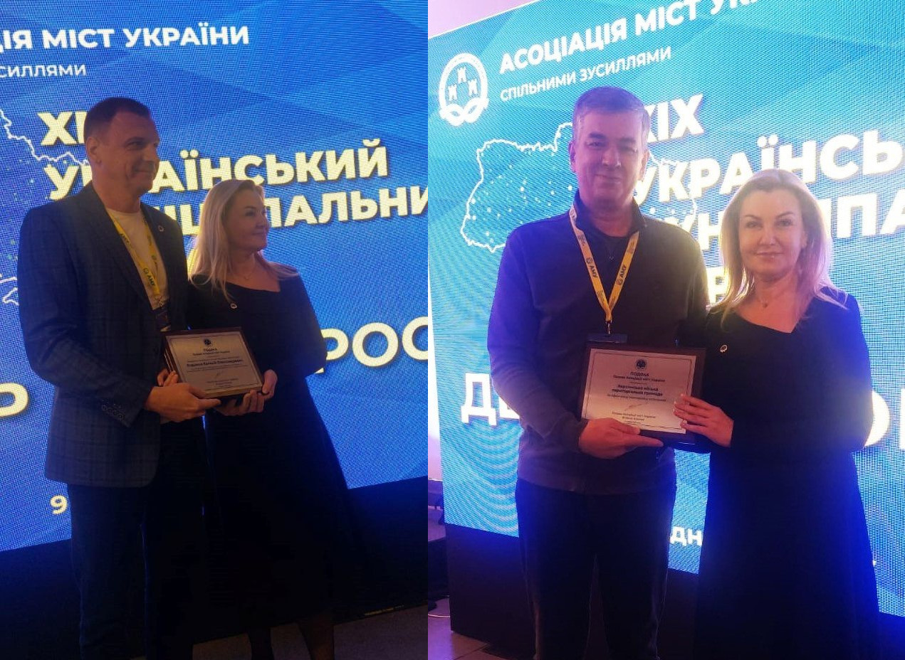 На банері з логотипом Асоціації міст України, на синьому фоні, дві окремі фотографії чоловіка з жінкою. На обох фото різні чоловіки і та сама жінка стоять поряд і тримають нагороду, дивлячись на камеру.
