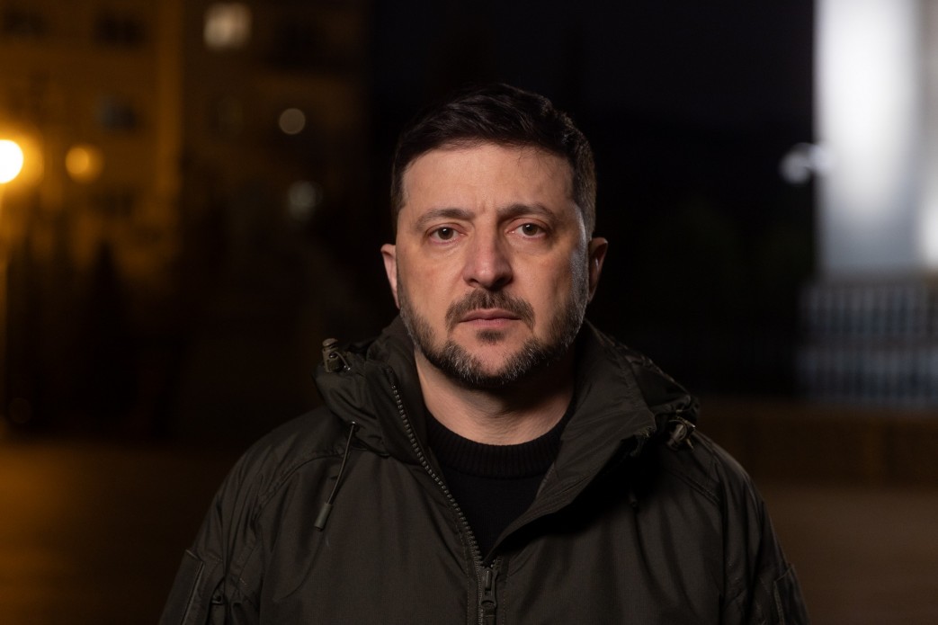 Чоловік середнього віку з короткою бородою дивиться прямо в камеру на фоні нічного міського пейзажу. Він одягнений у куртку кольору хакі з капюшоном, а м’яке освітлення підкреслює серйозний вираз його обличчя.