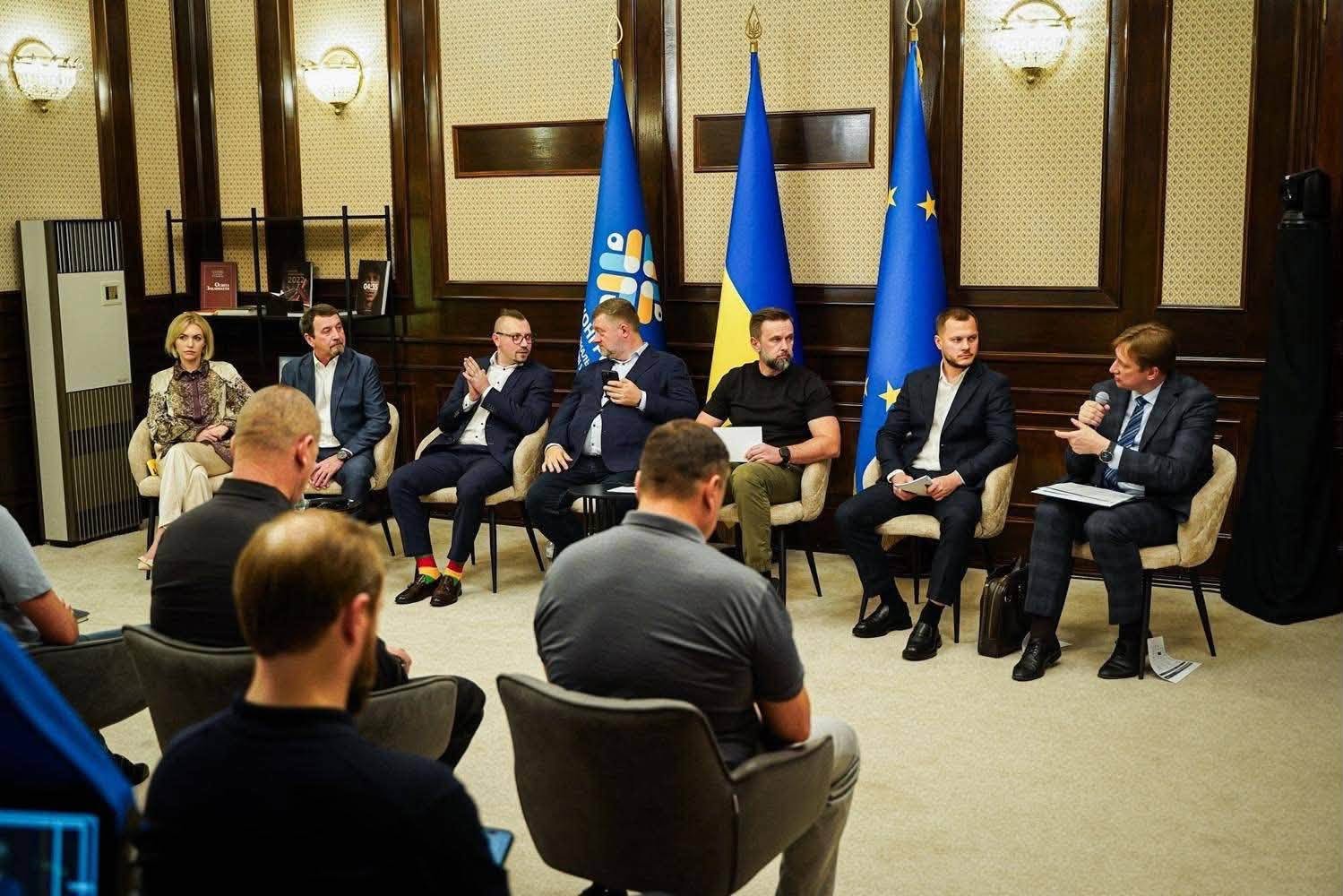Офіційне засідання у класично оздобленій залі з дерев’яними панелями. Спікери та президія сидять у півколі на м’яких кріслах. На задньому плані між ними встановлено три великі прапори: синій прапор Конгресу місцевих та регіональних влад, державний прапор України та прапор Європейського Союзу. Спікер праворуч говорить у мікрофон, у центрі сидить заступник Керівника Офісу Президента України Віктор Микита в чорній футболці, а перед ними розташовані стільці з учасниками наради.