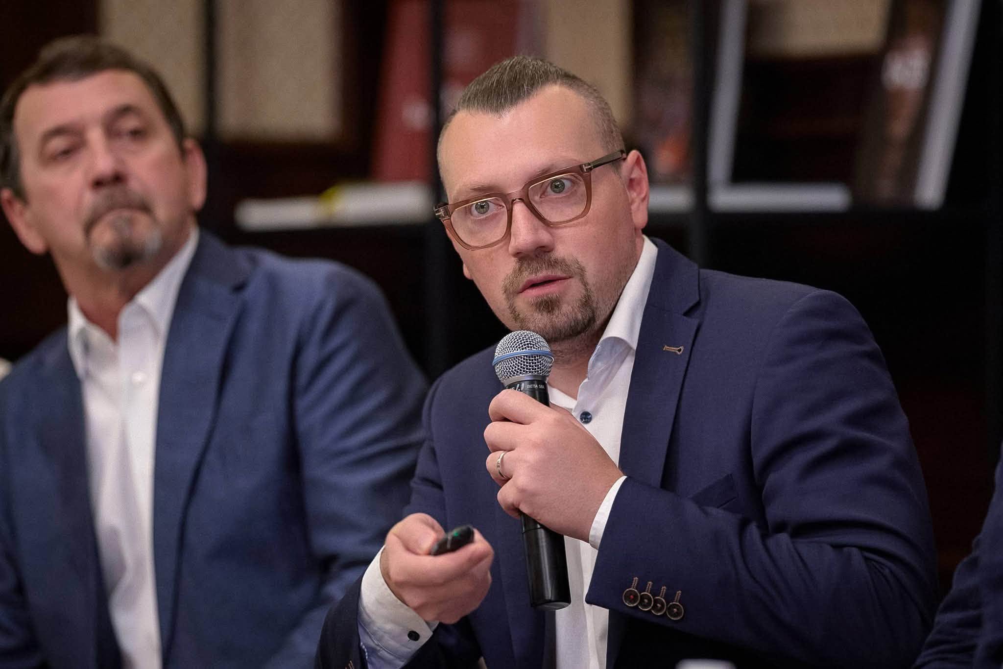 Крупний план міністра у справах громад, територій та внутрішньо переміщених осіб України Віталія Безгіна Чоловік говорить у мікрофон і тримає в іншій руці клікер для презентацій під час виступу. Ліворуч від нього за столом сидить інший колега у темно-синьому костюмі.