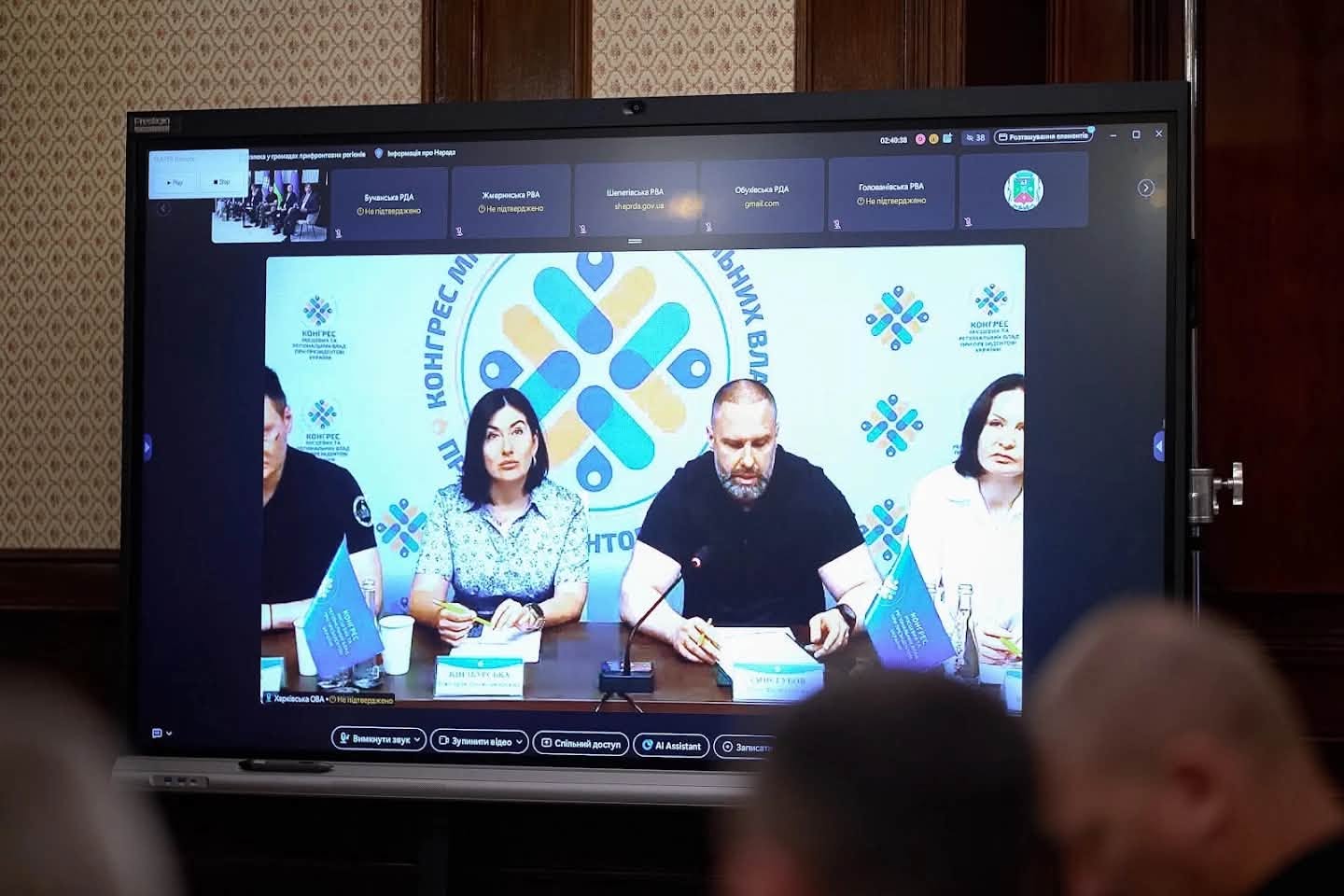 Великий монітор у залі засідань, на якому транслюється виступ спікерів у рамках засідання Конгресу місцевих та регіональних влад при Президентові України. У центрі екрана за столом із мікрофонами та табличками сидять четверо посадовців на тлі брендованого банера з логотипом Конгресу. На передньому плані видно силуети людей, які дивляться на екран.