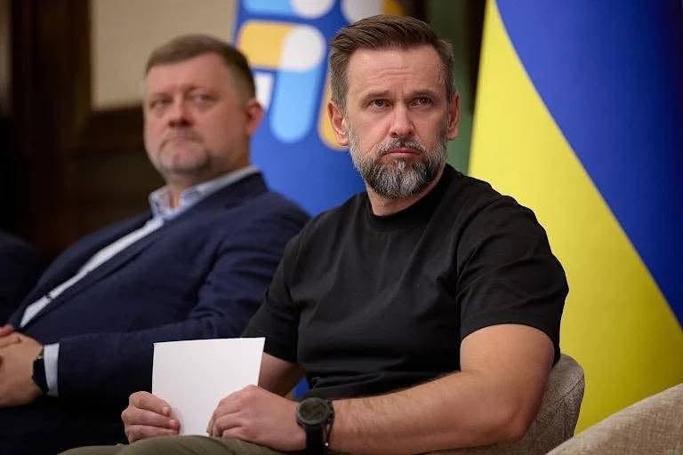 Портрет заступника Керівника Офісу Президента України Віктора Микити під час засідання. Він одягнений у чорну футболку, тримає в руках аркуш паперу та уважно слухає виступ колег. На задньому плані розмито видно державного прапора України та іншого учасника наради в темно-синьому піджаку.