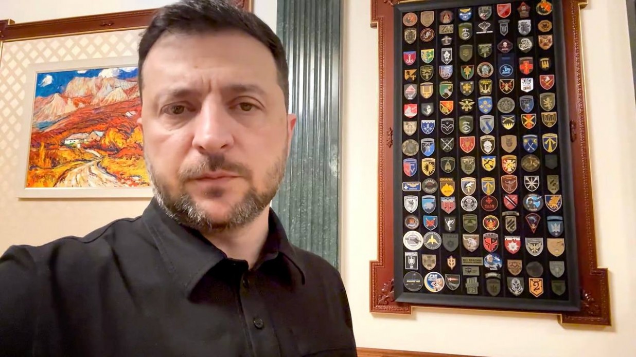 Президент України Володимир Зеленський у чорній сорочці дивиться прямо в камеру; на задньому плані видно картину та велику рамку з численними військовими шевронами.