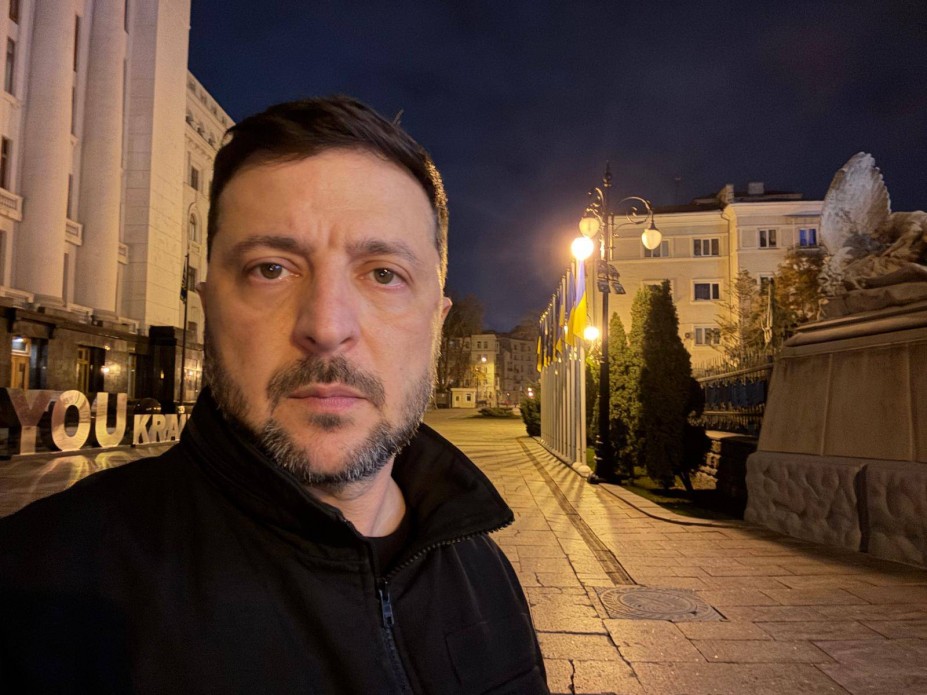 Портрет Президента України Володимира Зеленського в темній куртці, зроблений вночі на вулиці. На задньому плані видно мощену площу, адміністративну будівлю, а також ряд прапорів, що освітлені вуличними ліхтарями.