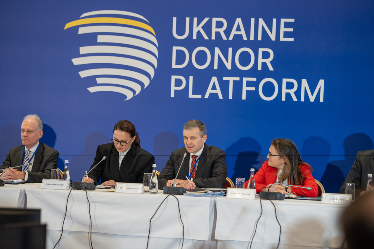 Четверо офіційних осіб у ділових костюмах сидять за столом під час засідання міжнародної платформи. На синьому фоні за ними розміщено напис «UKRAINE DONOR PLATFORM». Учасники роблять записи та обмінюються думками, перед ними встановлені мікрофони та таблички з іменами.