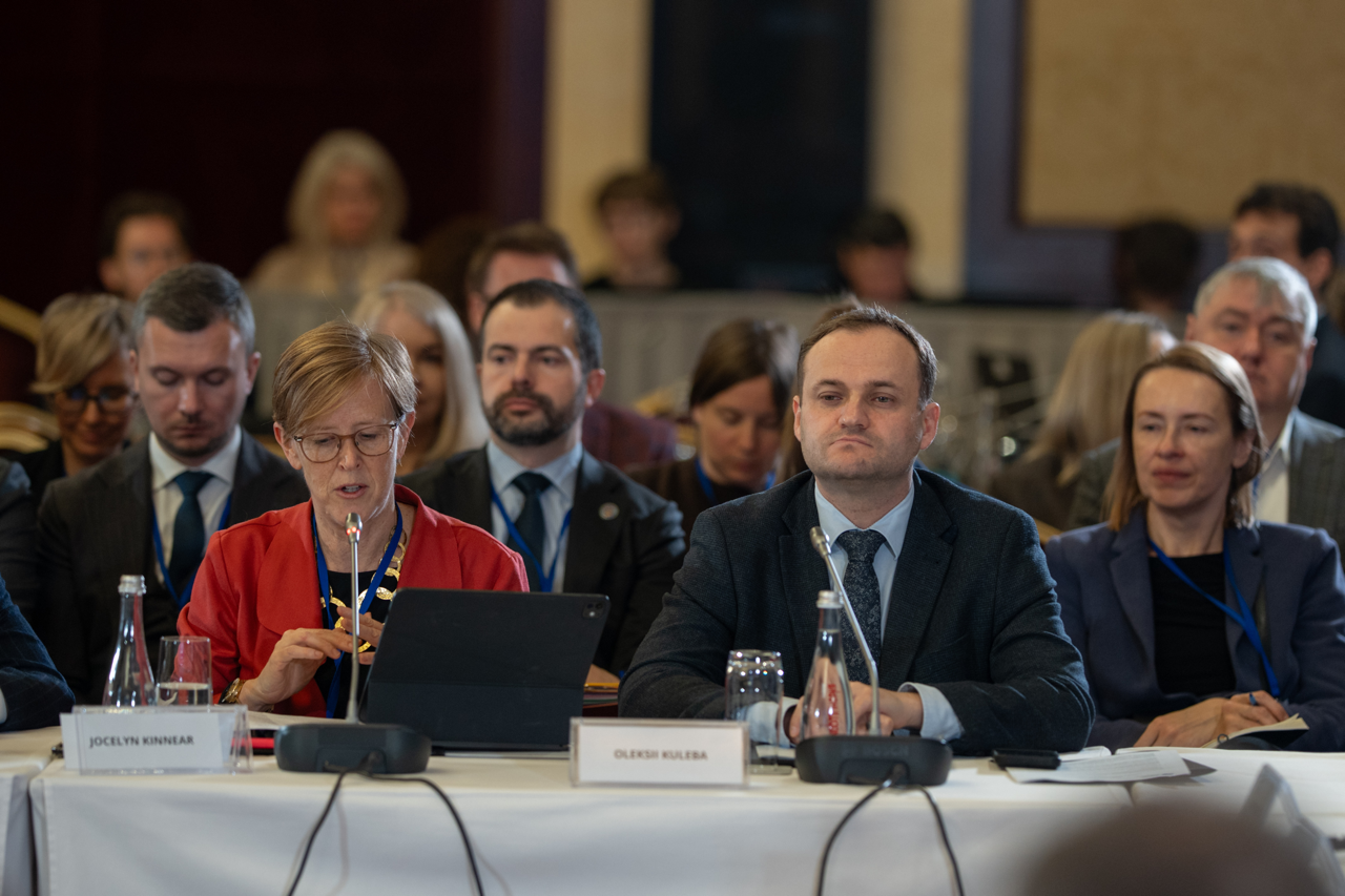 Група офіційних осіб у ділових костюмах сидить за довгим столом під час засідання. На передньому плані жінка в червоному піджаку працює з планшетом, а поруч із нею чоловік у темному костюмі зосереджено слухає виступ. На столі встановлені мікрофони та таблички з іменами учасників.