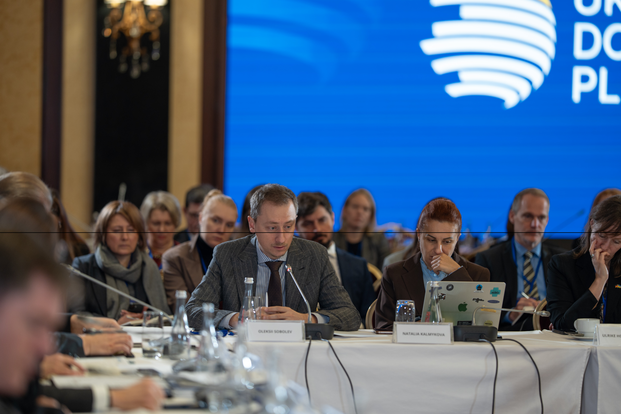 Чоловік у сірому піджаку та жінка в коричневому жакеті сидять за столом під час конференції. Жінка працює за ноутбуком, а на задньому плані видно великий синій екран з логотипом міжнародної донорської платформи та інших учасників заходу.