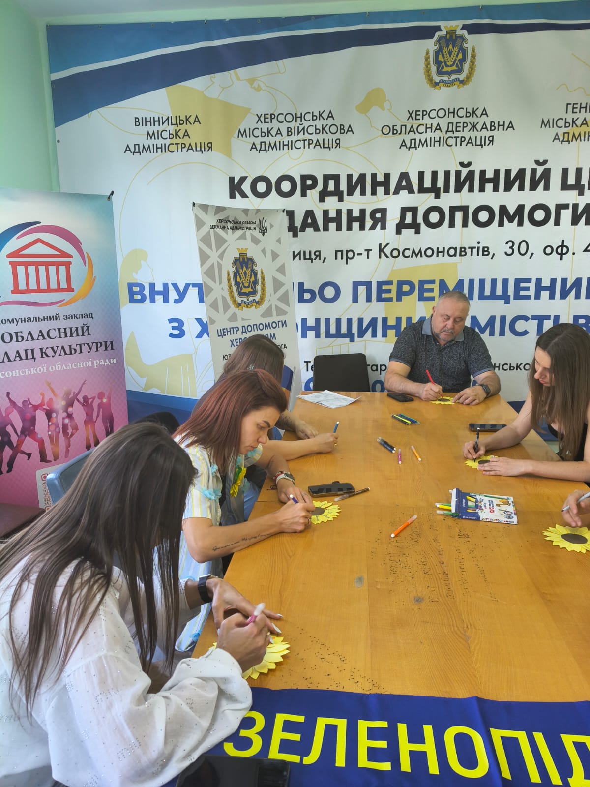 Працівники та відвідувачі координаційного центру допомоги ВПО з Херсонщини у місті Вінниця сидять за великим дерев'яним столом і фломастерами підписують паперові соняшники пам'яті. Позаду розміщено великі інформаційні банери Вінницької та Херсонської міських військових адміністрацій, Херсонської ОДА та Обласного палацу культури, а на передньому плані видно край синьо-жовтого прапора Зеленопідської громади.