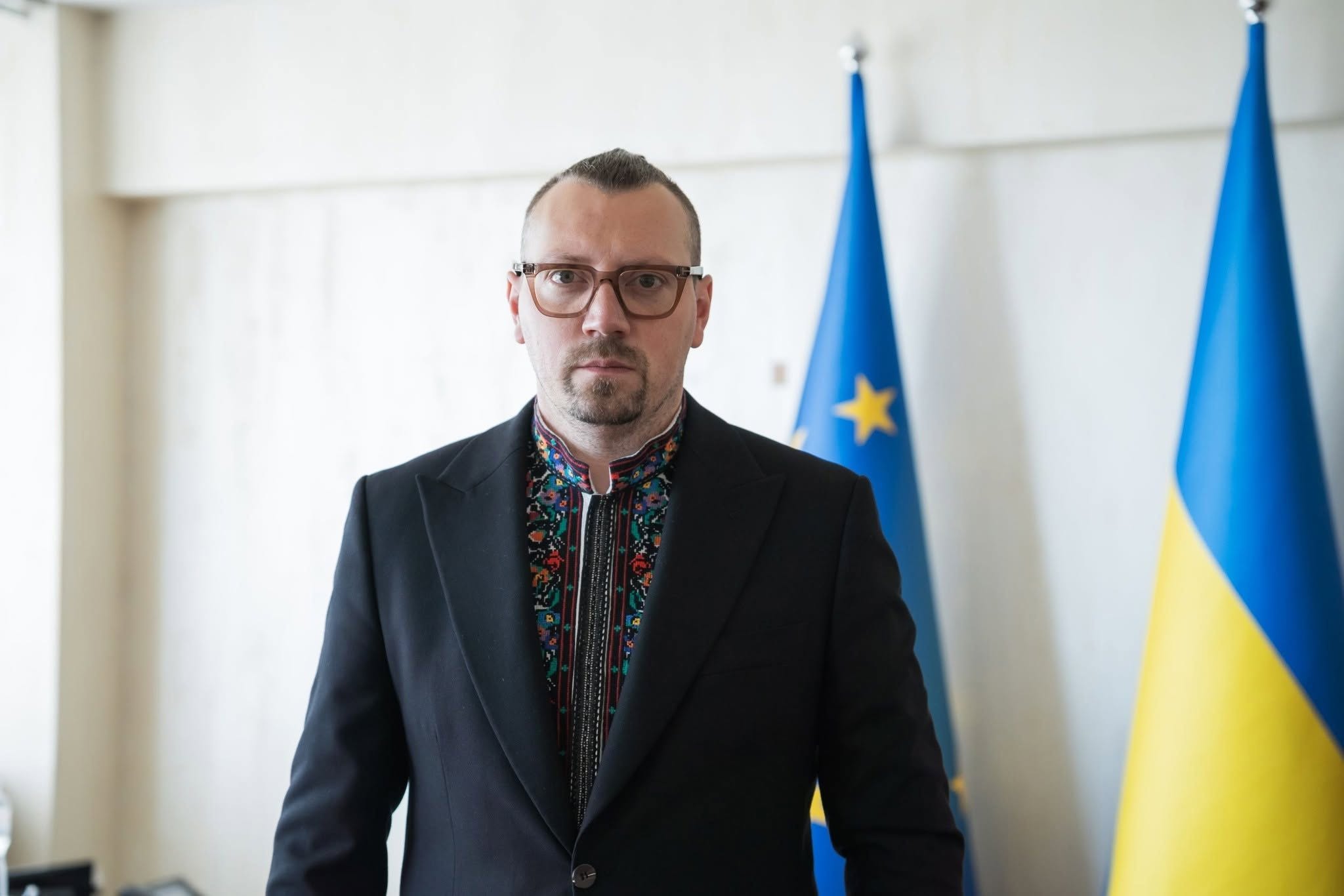 Чоловік в окулярах у масивній коричневій оправі та з акуратною борідкою дивиться прямо в камеру. Він одягнений у чорний класичний піджак поверх темної вишиванки з геометрично-рослинним орнаментом на комірі та планці.  На розмитому задньому плані видно світлу стіну кабінету та два встановлені поруч прапори на флагштоках: прапор Європейського Союзу та синьо-жовтий Державний Прапор України.