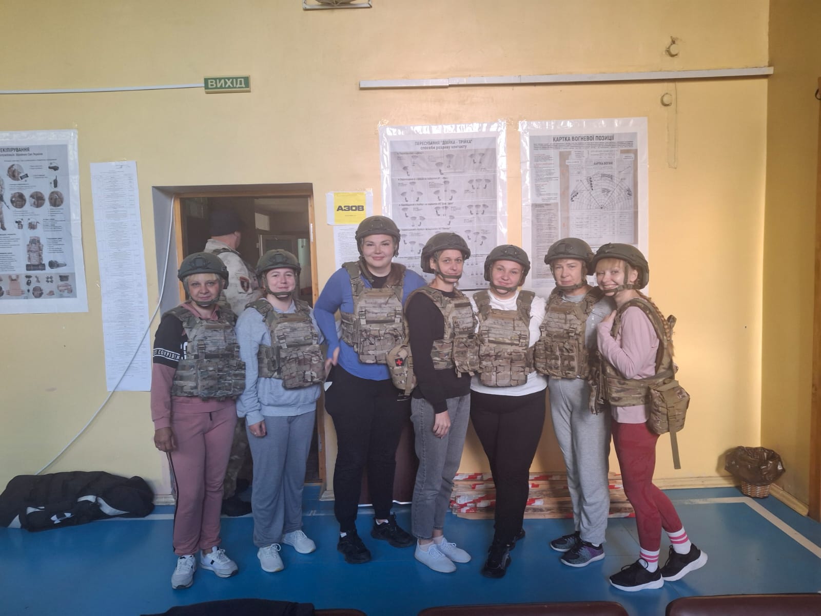 Група із семи цивільних жінок стоїть у ряд у навчальному приміщенні під час проходження тренінгу з безпеки або тактичної підготовки.  Усі учасниці екіпіровані в захисні шоломи (каски) та бронежилети камуфляжного забарвлення мультикам і піксель поверх повсякденного спортивного одягу. На стіні позаду висять навчальні інформаційні плакати зі схемами бойових порядків («Пересування двійка-трійка», «Картка вогневої позиції»), плакат про індивідуальне екіпірування військовослужбовців та жовта табличка «АЗОВ».