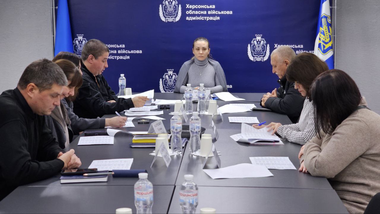 Група людей сидить за великим столом під час наради чи зустрічі. Жінка в центрі дивиться прямо, інші зосереджені на документах. На стіні позаду видно напис: Херсонська обласна військова адміністрація.