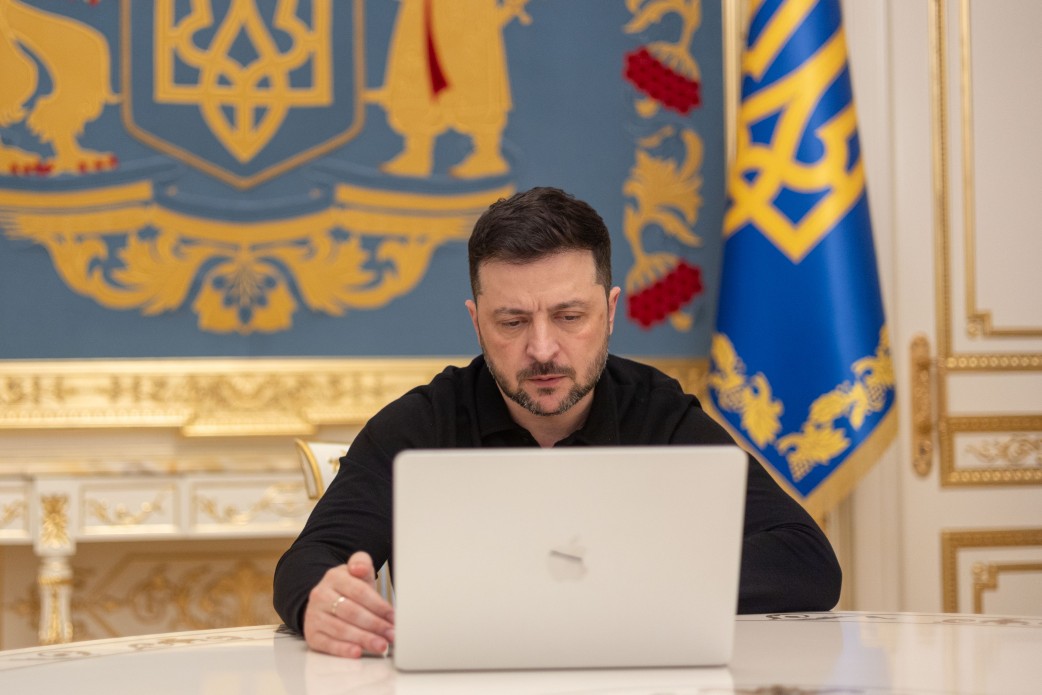 Володимир Зеленський, Президент України, сидить за столом у кабінеті, дивлячись на відкритий ноутбук Apple. На задньому плані розташований великий синій стілець з золотим Тризубом (Гербом України) та синьо-жовтий прапор.