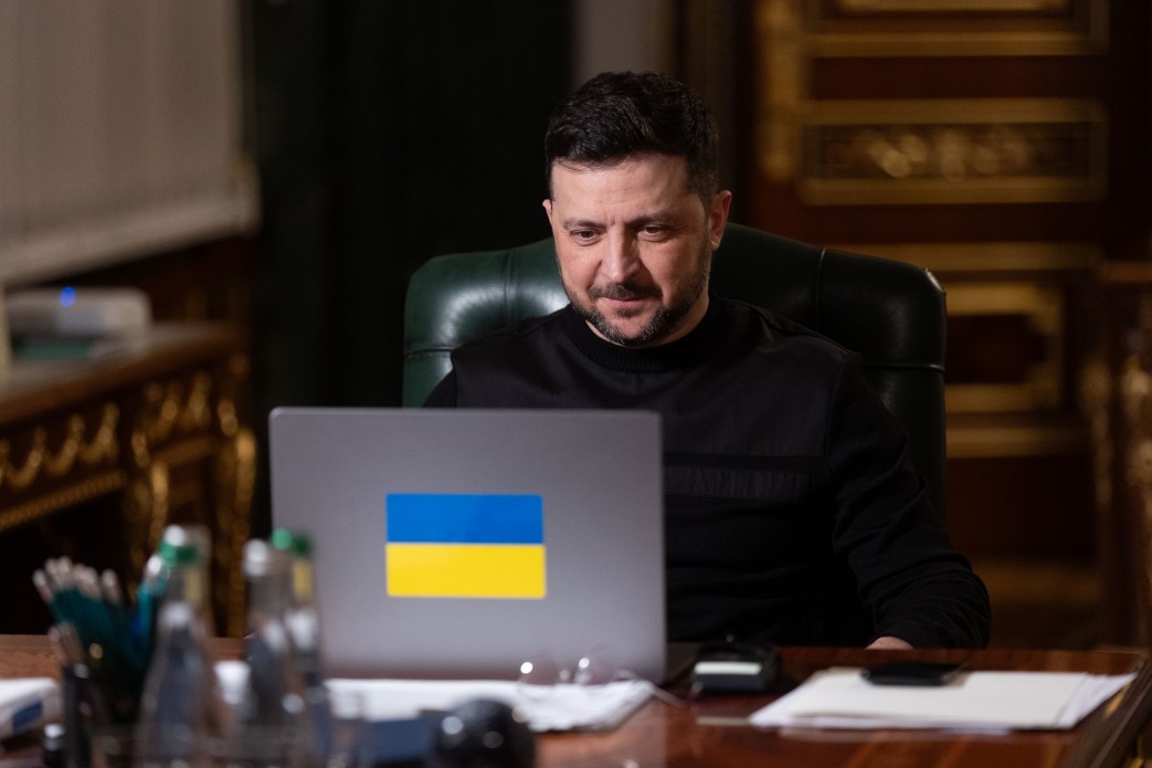 Чоловік у чорному светрі сидить за робочим столом і зосереджено дивиться в ноутбук, на кришці якого зображений прапор України.