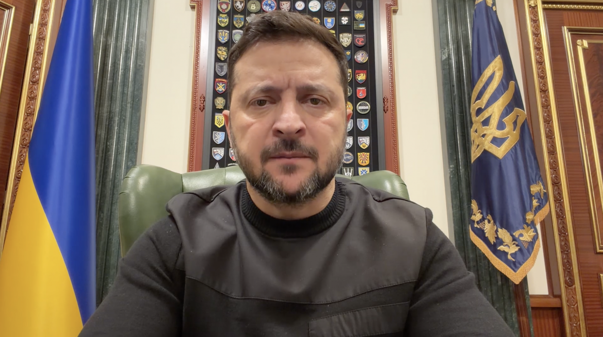 Чоловік у чорному одязі звертається до глядачів на фоні державного прапора та штандарта з тризубом. Позаду нього на стіні розміщена панель із військовими шевронами.