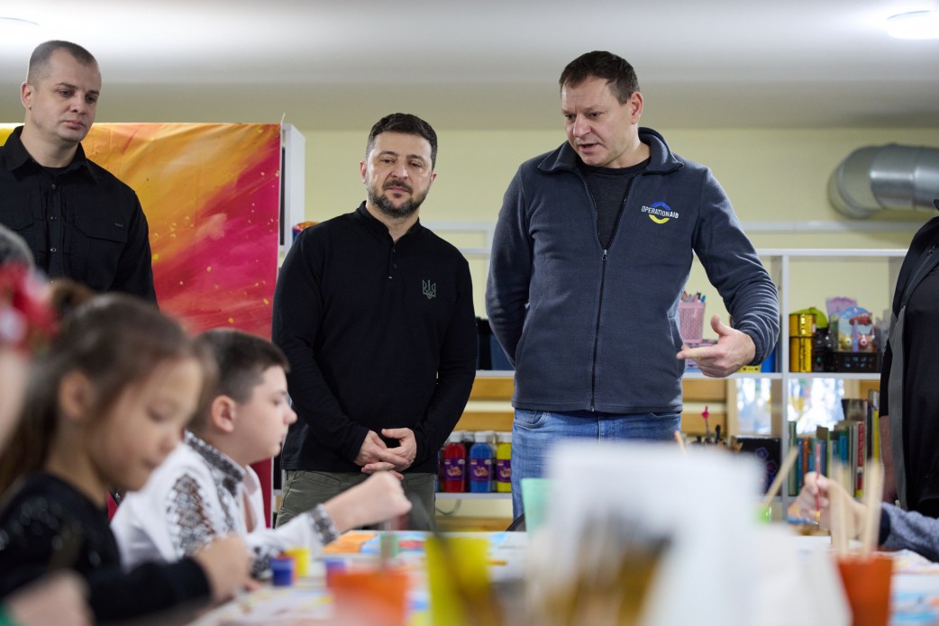 Президент України Володимир Зеленський (в чорній кофті з тризубом) стоїть у дитячому приміщенні поруч із чоловіком у темно-сірій кофті на блискавці, який щось пояснює. На передньому плані діти зайняті малюванням за столом.