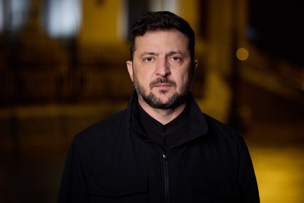 Крупний план-портрет Володимира Зеленського, Президента України, знятий на вулиці вночі. Він дивиться прямо у камеру із серйозним, зосередженим виразом обличчя. У нього коротка темна стрижка та акуратна борода. На ньому чорна куртка на блискавці з коміром-стійкою та накладними кишенями, одягнена поверх чорної сорочки.