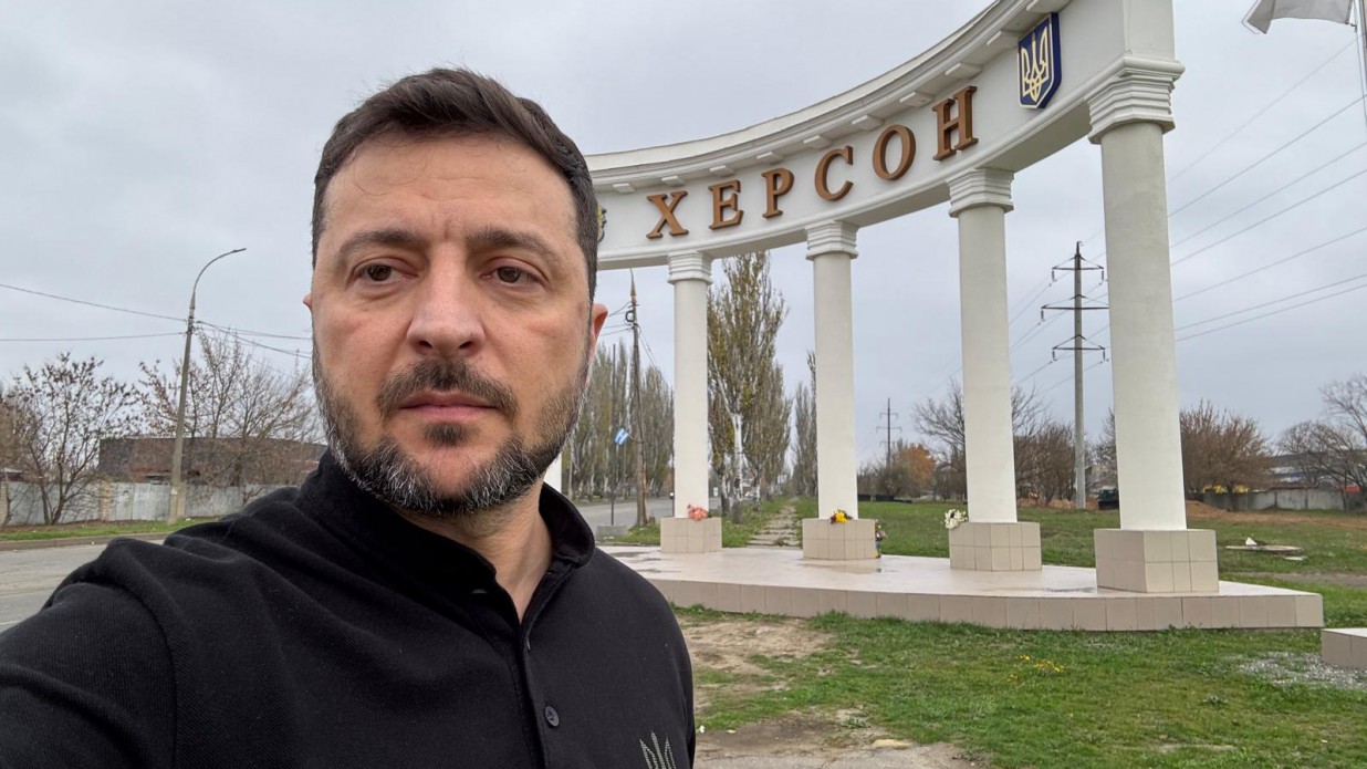 Селфі Президента України Володимира Зеленського на тлі стели з написом: Херсон та гербом України, розташованої на в'їзді в місто. Президент одягнений у чорну кофту. Погода похмура, осіння.