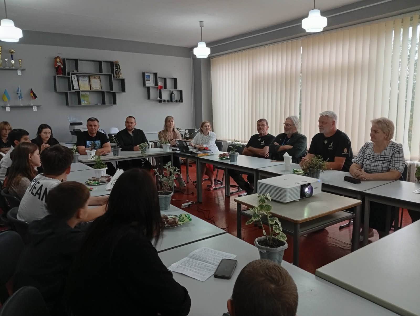 Загальний план круглого столу в просторому кабінеті зі світлими жалюзі на великих вікнах. Учасники заходу — діти, молодь і дорослі іноземні гості та представники громади — сидять навколо зсунутих столів у формі літери «П». На окремому столику в центрі встановлено білий мультимедійний проєктор, на столах стоять кімнатні квіти в горщиках, частування та робочі матеріали.