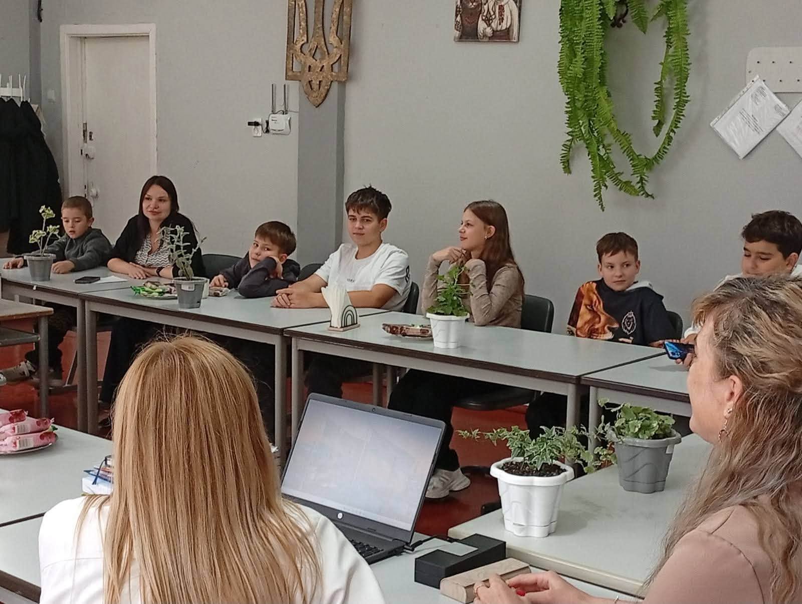 Діти та підлітки сидять за партами вздовж стіни кабінету під час зустрічі або інтерактивного уроку. На передньому плані зі спини видно двох ведучих перед відкритим ноутбуком. На столах розставлені невеликі кімнатні рослини у вазонах і серветки. Позаду на сірій стіні закріплено дерев'яний різьблений герб України — Тризуб, картину та довгі гілки декоративної зеленої папороті.