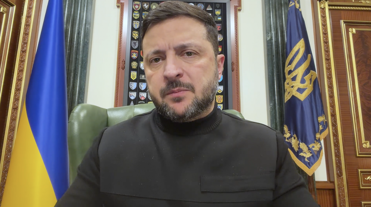 Чоловік у темному одязі виступає з промовою на фоні державного прапора України та штандарта Президента України. За ним розташований стенд із військовими шевронами та емблемами різних підрозділів.