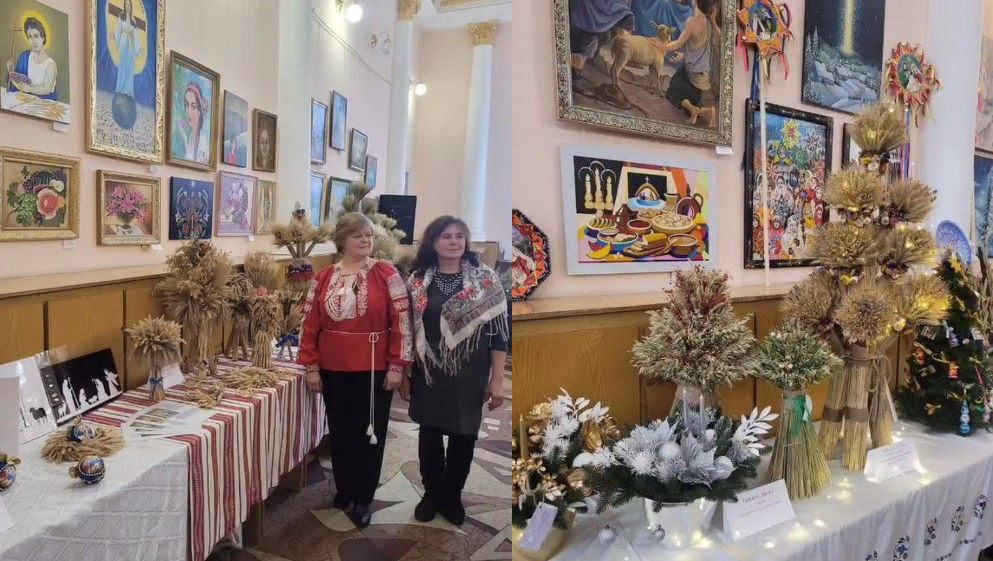 Дві жінки, одягнені у вишиванку (червона) та хустку (сіра), стоять у виставковому залі. Перед ними розташований довгий стіл, накритий скатертиною в червоно-білу смужку, на якому представлені численні декоративні вироби. Це букети та композиції із золотистих колосків пшениці та інших сухоцвітів, а також стилізовані ялинки та менші букети, прикрашені білими та сріблястими елементами. На задньому плані стіни прикрашені великою кількістю картин.