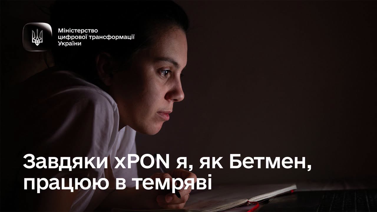 Альтернативний текстІнформаційний банер Міністерства цифрової трансформації України про енергонезалежний інтернет за технологією xPON.   На темному фоні зображено дівчину в білій футболці в темній кімнаті, обличчя якої підсвічується холодним світлом від екрана ноутбука. Вона зосереджено дивиться вперед, тримаючи в руці ручку біля розгорнутого блокнота на столі.   У лівому верхньому кутку розміщено малий Державний Герб України та напис: «Міністерство цифрової трансформації України».   У нижній частині великими білими літерами нанесено текст: «Завдяки xPON я, як Бетмен, працюю в темряві».   
