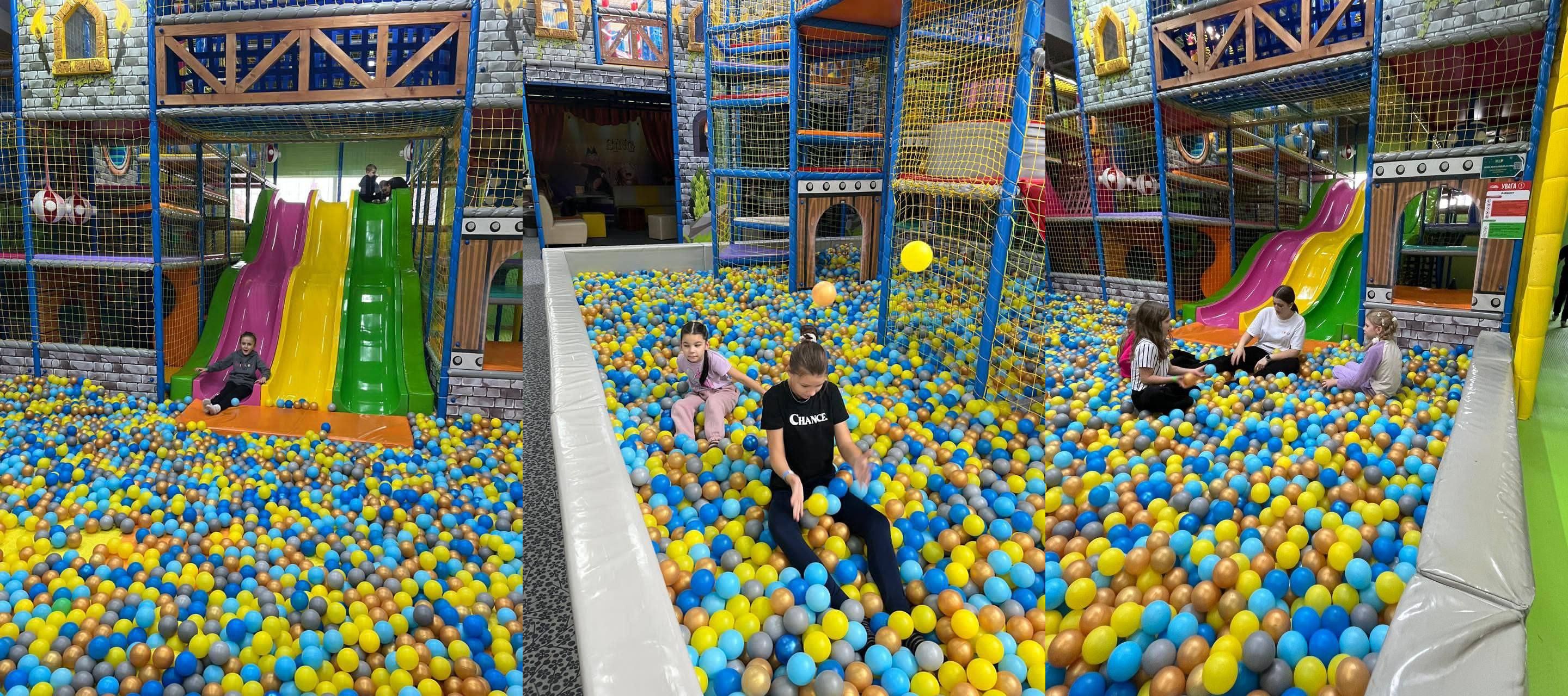 Панорамний знімок великої ігрової зони у розважальному центрі, де діти граються у величезному басейні, наповненому синіми, жовтими та золотистими кульками. На тлі видно високі ігрові конструкції у вигляді замку з гірками та сітками.