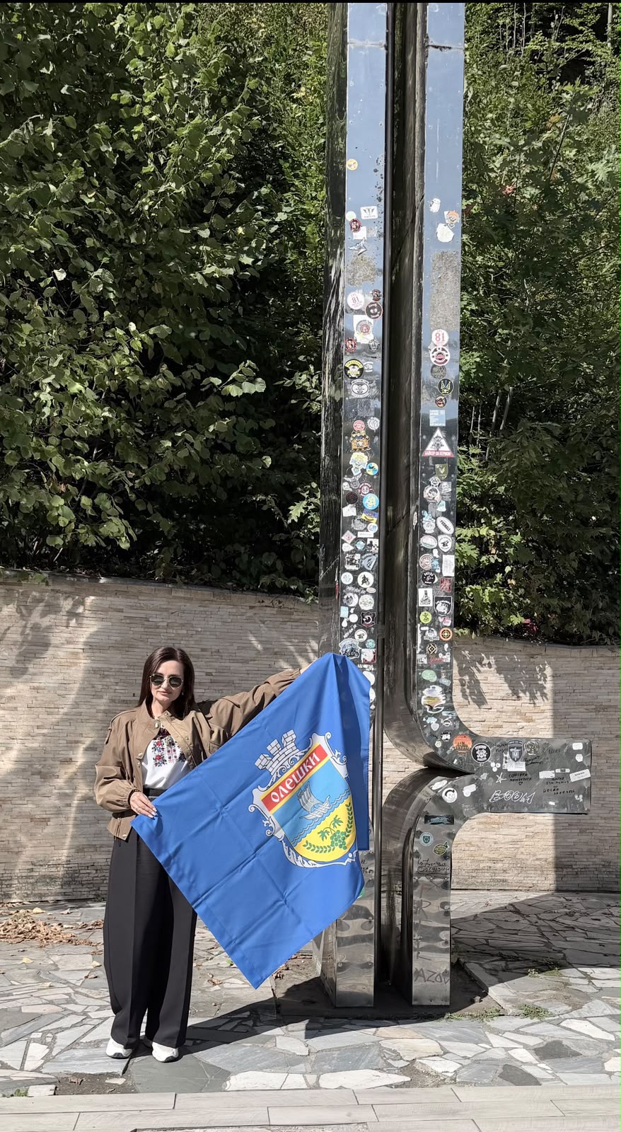 Жінка в сонцезахисних окулярах, бежевій куртці, вишиванці та чорних палаццо стоїть біля високої дзеркальної металевої стели, густо обклеєної пам'ятними та патріотичними наліпками. Вона розгортає перед собою синій прапор міста Олешки з міським гербом і підписом «ОЛЕШКИ». Фото зроблене на тлі кам'яної стіни та зелених дерев.