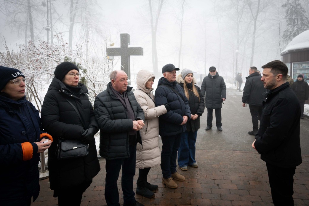 Чоловік у темному верхньому одязі спілкується з групою людей у зимовому вбранні на відкритому повітрі. Зустріч відбувається у засніженому парку, на задньому плані серед дерев видніється великий кам'яний хрест. Учасники розмови виглядають зосередженими та серйозними.